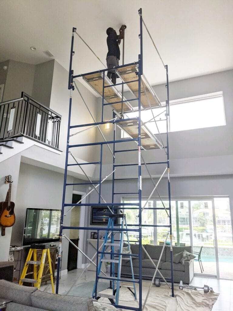 A worker installing or repairing electrical fixtures on a high ceiling inside a modern living room with large windows and natural light.