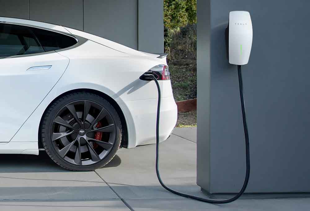 White electric vehicle parked in a home garage, connected to a hardwired EV charger mounted on the wall. The charging cord is securely plugged into the car's charging port, illustrating the setup of a permanent home charging solution.