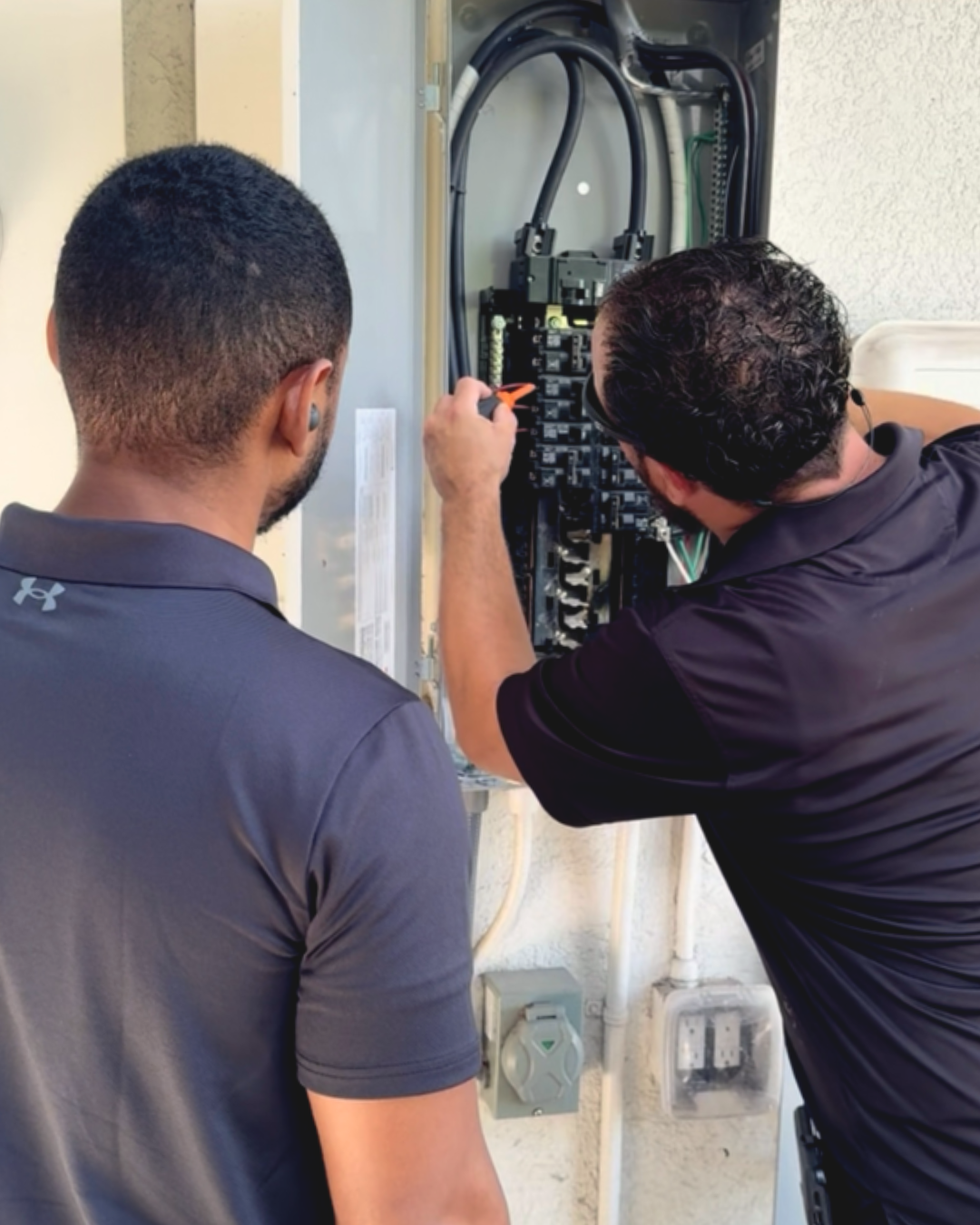 Two men wearing black collared shirts working on electrical panel
